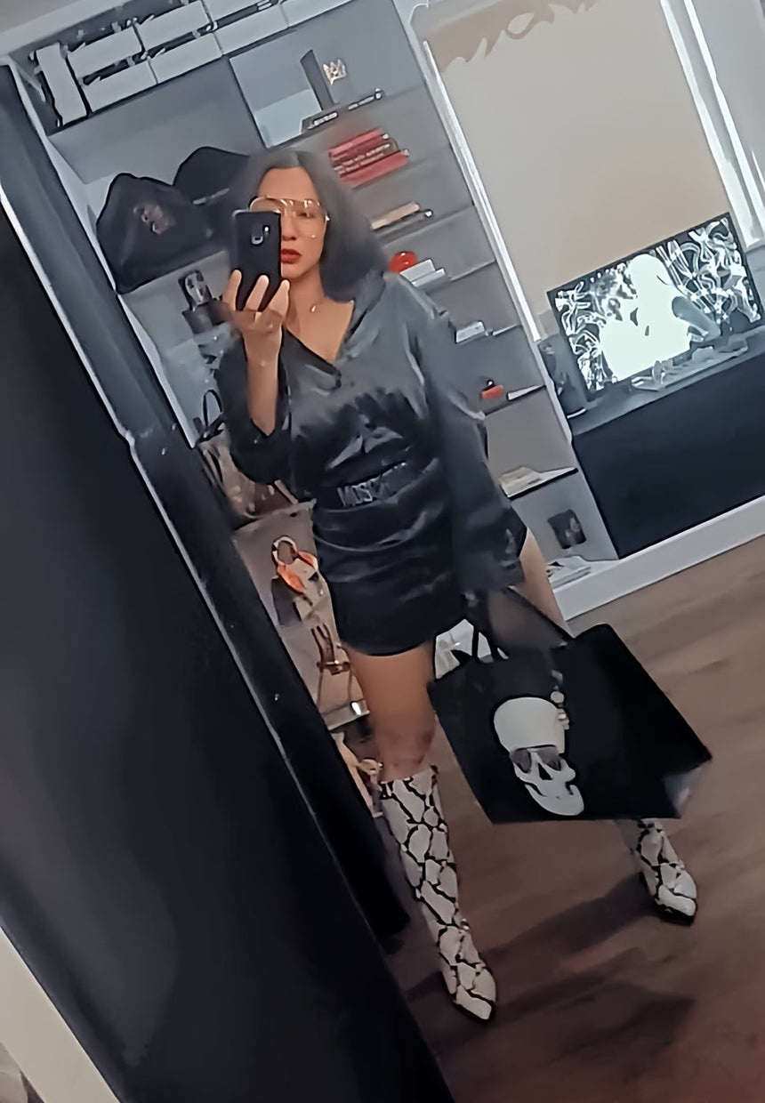 Person taking a mirror selfie wearing a black dress, white boots, and holding a black bag with a skull design.