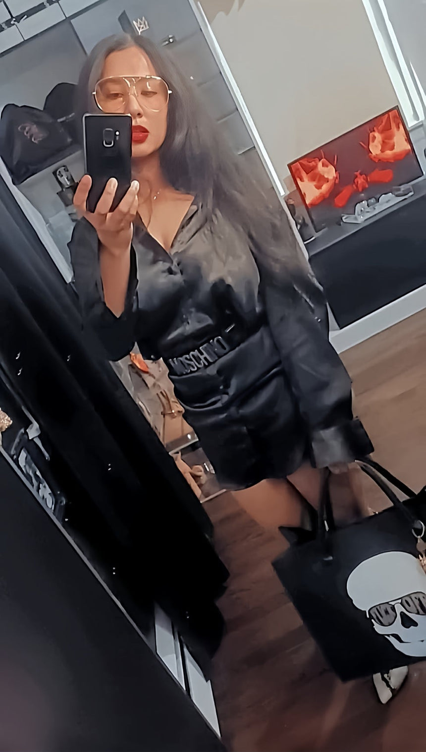 Person taking a mirror selfie wearing a black outfit with text, holding a black bag with a skull design.
